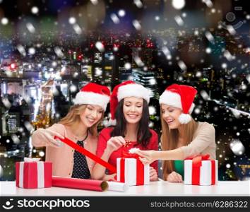 christmas, holidays, celebration, decoration and people concept - smiling women in santa helper hats with decorating paper and gift boxes over snowy night city background