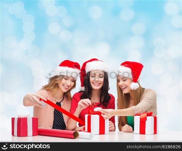 christmas, holidays, celebration, decoration and people concept - smiling women in santa helper hats with decorating paper and gift boxes over blue lights background