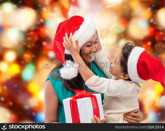 christmas, holidays, celebration, family and people concept - happy mother and child girl in santa helper hats with gift box over red lights background