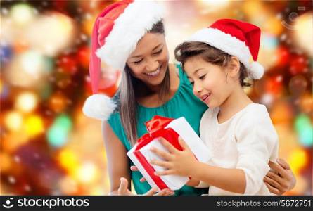 christmas, holidays, celebration, family and people concept - happy mother and little girl in santa helper hats with gift box over red lights background