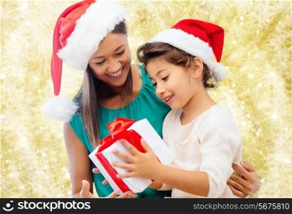christmas, holidays, celebration, family and people concept - happy mother and little girl in santa helper hats with gift box over yellow lights background