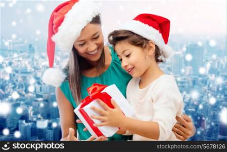 christmas, holidays, celebration, family and people concept - happy mother and little girl in santa helper hats with gift box over snowy city background