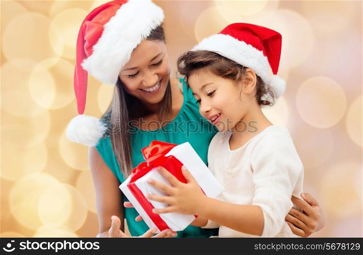 christmas, holidays, celebration, family and people concept - happy mother and little girl in santa helper hats with gift box over beige lights background