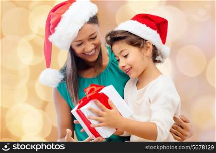 christmas, holidays, celebration, family and people concept - happy mother and little girl in santa helper hats with gift box over beige lights background