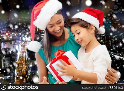 christmas, holidays, celebration, family and people concept - happy mother and little girl in santa helper hats with gift box over snowy night city background