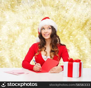 christmas, holidays, celebration, greeting and people concept - smiling woman in santa helper hat with gift box writing letter or sending post card over yellow lights background