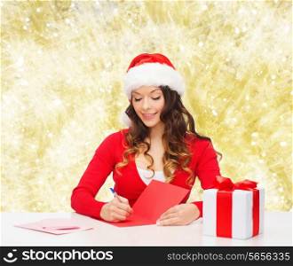 christmas, holidays, celebration, greeting and people concept - smiling woman in santa helper hat with gift box writing letter or sending post card over yellow lights background