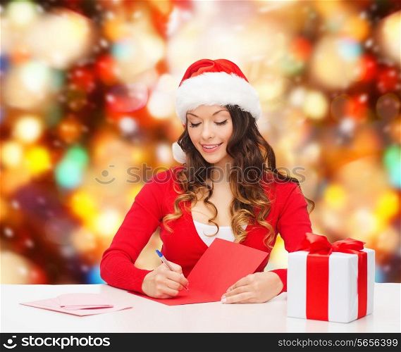 christmas, holidays, celebration, greeting and people concept - smiling woman in santa helper hat with gift box writing letter or sending post card over red lights background