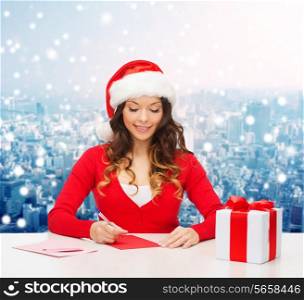 christmas, holidays, celebration, greeting and people concept - smiling woman in santa helper hat with gift box writing letter or sending post card over snowy city background