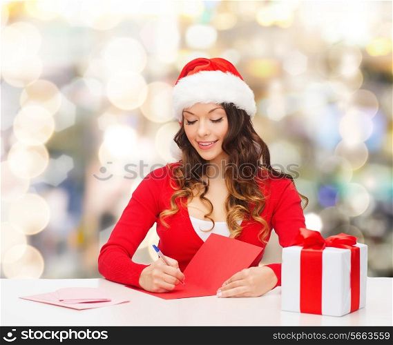 christmas, holidays, celebration, greeting and people concept - smiling woman in santa helper hat with gift box writing letter or sending post card over lights background