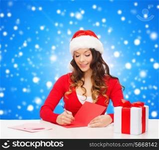 christmas, holidays, celebration, greeting and people concept - smiling woman in santa helper hat with gift box writing letter or sending post card over blue snowy background