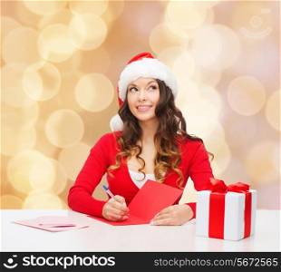 christmas, holidays, celebration, greeting and people concept - smiling woman in santa helper hat with gift box writing letter or sending post card over beige lights background