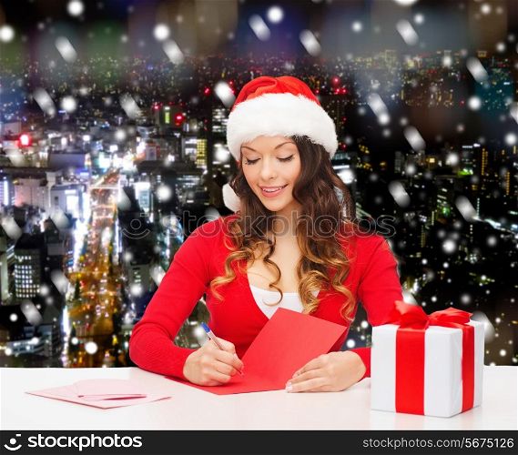 christmas, holidays, celebration, greeting and people concept - smiling woman in santa helper hat with gift box writing letter or sending post card over snowy night city background