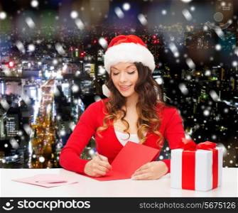 christmas, holidays, celebration, greeting and people concept - smiling woman in santa helper hat with gift box writing letter or sending post card over snowy night city background