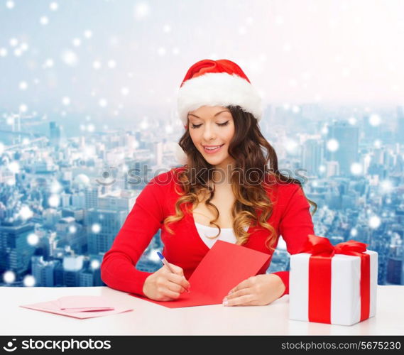 christmas, holidays, celebration, greeting and people concept - smiling woman in santa helper hat with gift box writing letter or sending post card over snowy city background