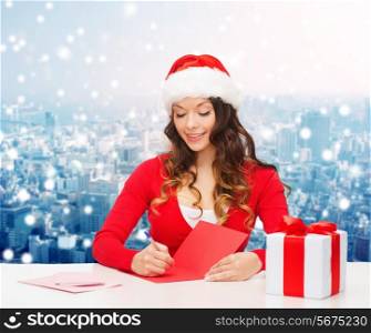 christmas, holidays, celebration, greeting and people concept - smiling woman in santa helper hat with gift box writing letter or sending post card over snowy city background