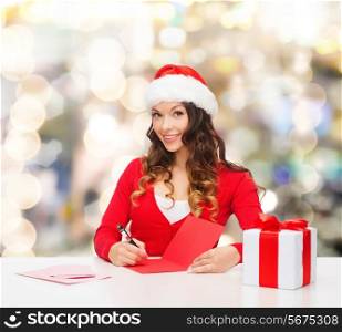 christmas, holidays, celebration, greeting and people concept - smiling woman in santa helper hat with gift box writing letter or sending post card over lights background
