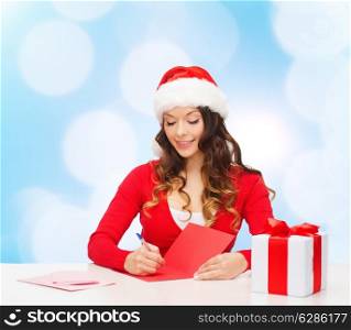 christmas, holidays, celebration, greeting and people concept - smiling woman in santa helper hat with gift box writing letter or sending post card over blue lights background