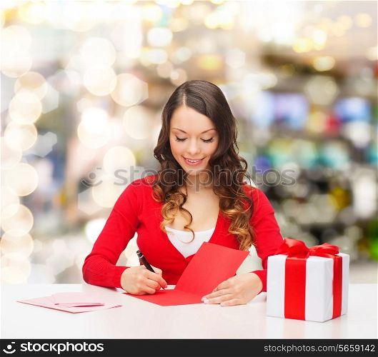 christmas, holidays, celebration, greeting and people concept - smiling woman with gift box writing letter or sending post card over lights background