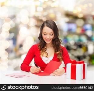 christmas, holidays, celebration, greeting and people concept - smiling woman with gift box writing letter or sending post card over lights background