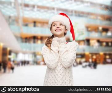christmas, holidays, childhood and people concept - smiling girl in santa helper hat over shopping center background