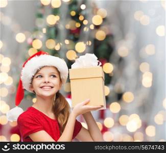 christmas, holidays, childhood and people concept - smiling girl in santa helper hat with gift box over christmas tree lights background
