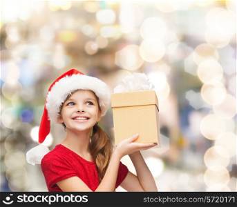 christmas, holidays, childhood and people concept - smiling girl in santa helper hat with gift box over lights background
