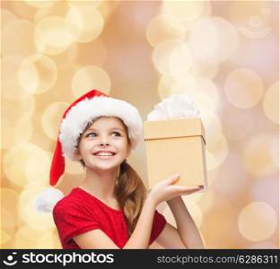 christmas, holidays, childhood and people concept - smiling girl in santa helper hat with gift box over beige lights background