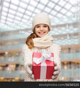 christmas, holidays, childhood, presents and people concept - dreaming girl in winter clothes with gift box over shopping center background