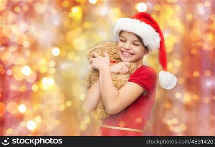 christmas, holidays, children and happiness concept - smiling girl in santa helper hat with teddy bear over lights background