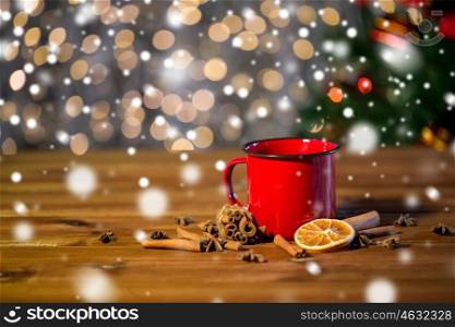 christmas, holidays, cooking and spice concept - close up of tea cup with cinnamon, anise and dried orange on wooden table over lights