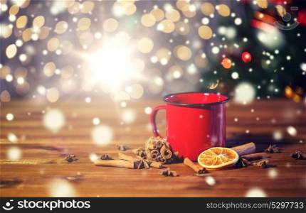 christmas, holidays, cooking and spice concept - close up of tea cup with cinnamon, anise and dried orange on wooden table over lights. tea cup with winter spices on wooden table