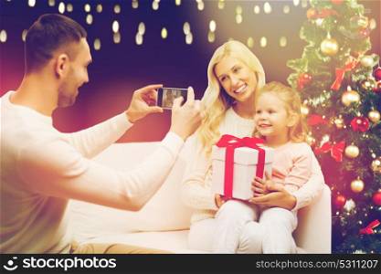 christmas, holidays, family and people concept - father photographing happy mother and little daughter with present by smartphone over lights background. man photographing his family with christmas gift