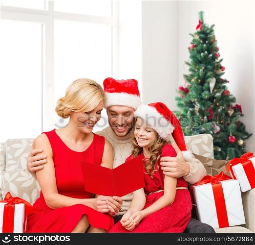 christmas, holidays, family and people concept - happy mother, father and little girl in santa helper hats with gift boxes reading geeting card over living room and christmas tree background