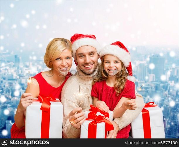 christmas, holidays, family and people concept - happy mother, father and little girl in santa helper hats with gift boxes and sparklers over snowy city background