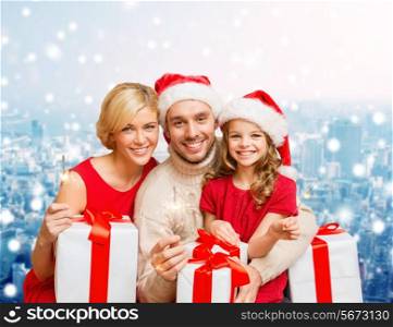 christmas, holidays, family and people concept - happy mother, father and little girl in santa helper hats with gift boxes and sparklers over snowy city background