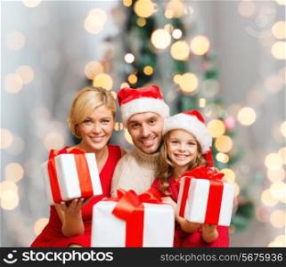 christmas, holidays, family and people concept - happy mother, father and little girl in santa helper hats with gift boxes over living room and christmas tree background