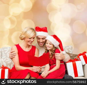 christmas, holidays, family and people concept - happy mother, father and little girl in santa helper hats with gift boxes reading geeting card over beige lights background