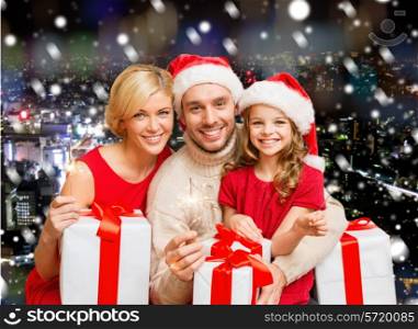 christmas, holidays, family and people concept - happy mother, father and little girl in santa helper hats with gift boxes and sparklers over snowy night city background