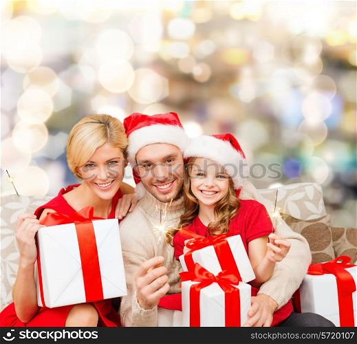 christmas, holidays, family and people concept - happy mother, father and little girl in santa helper hats with gift boxes and sparklers over lights background
