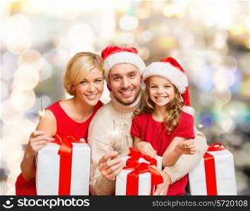 christmas, holidays, family and people concept - happy mother, father and little girl in santa helper hats with gift boxes and sparklers over lights background