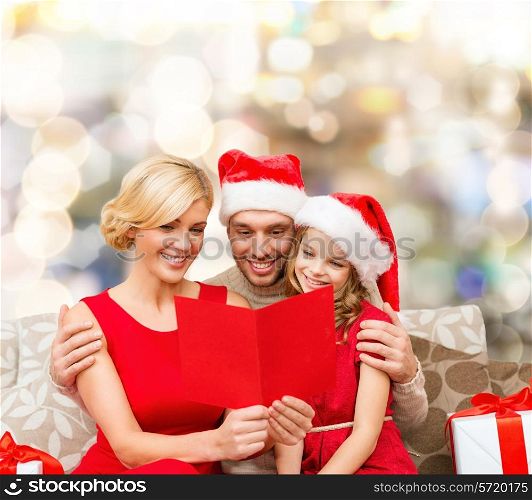 christmas, holidays, family and people concept - happy mother, father and little girl in santa helper hats with gift boxes reading geeting card over lights background