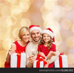christmas, holidays, family and people concept - happy mother, father and little girl in santa helper hats with gift boxes and sparklers over beige lights background