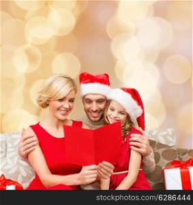christmas, holidays, family and people concept - happy mother, father and little girl in santa helper hats with gift boxes reading geeting card over beige lights background