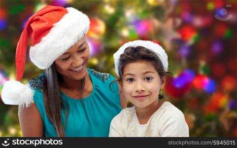 christmas, holidays, family, childhood and people concept - happy mother and little girl in santa hats over lights background