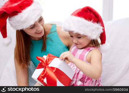 christmas, holidays, family, childhood and people concept - happy mother and little girl in santa hats with gift box at home