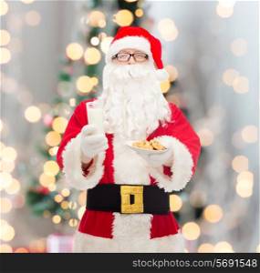 christmas, holidays, food, drink and people concept - man in costume of santa claus with glass of milk and cookies over tree lights background