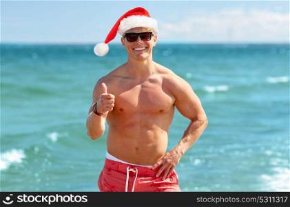 christmas, holidays, gesture and people concept - happy smiling young man in santa hat showing thumbs up on beach over sea. happy man showing thumbs up on beach at christmas
