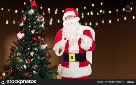 christmas, holidays, gesture and people concept - man in costume of santa claus with bag and christmas tree showing thumbs up over garland lights background. santa claus with bag and christmas tree
