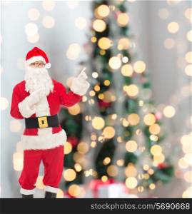 christmas, holidays, gesture and people concept - man in costume of santa claus pointing fingers over tree lights background